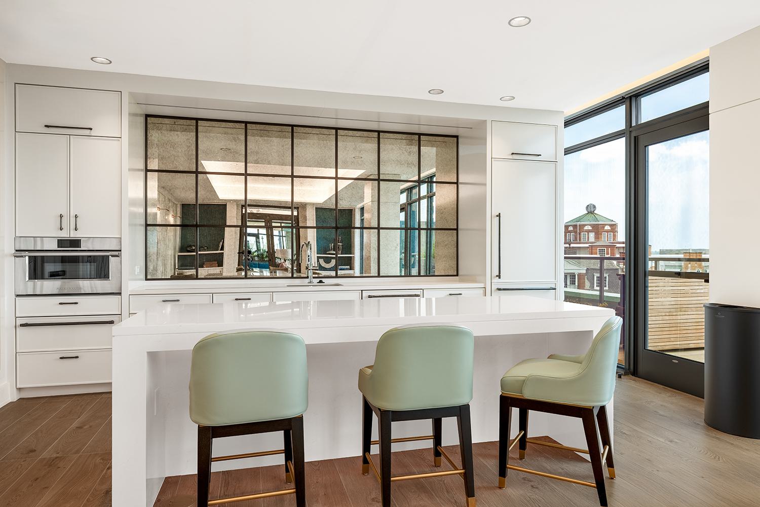 Entertaining kitchen at Aerie with a long white island, three mint-green upholstered stools, full-height white cabinetry, built-in appliances, and a large gridded mirror reflecting the adjacent lounge.