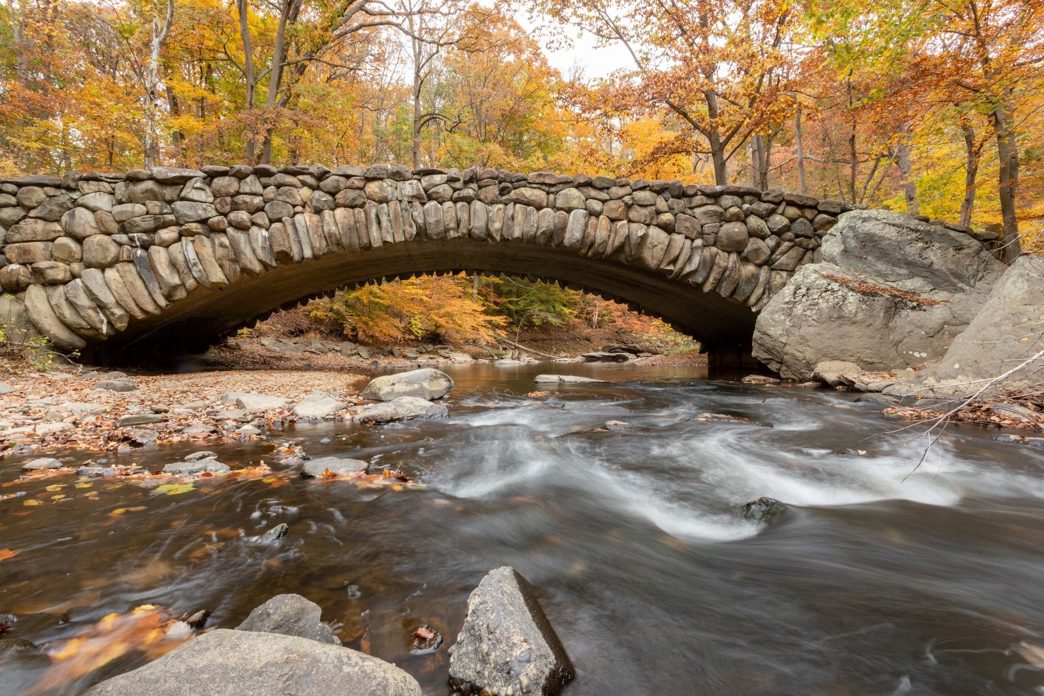 Boulder Bridge in Rock Creek Park, a historic stone arch spanning a shallow, fast-moving creek framed by colorful autumn trees.