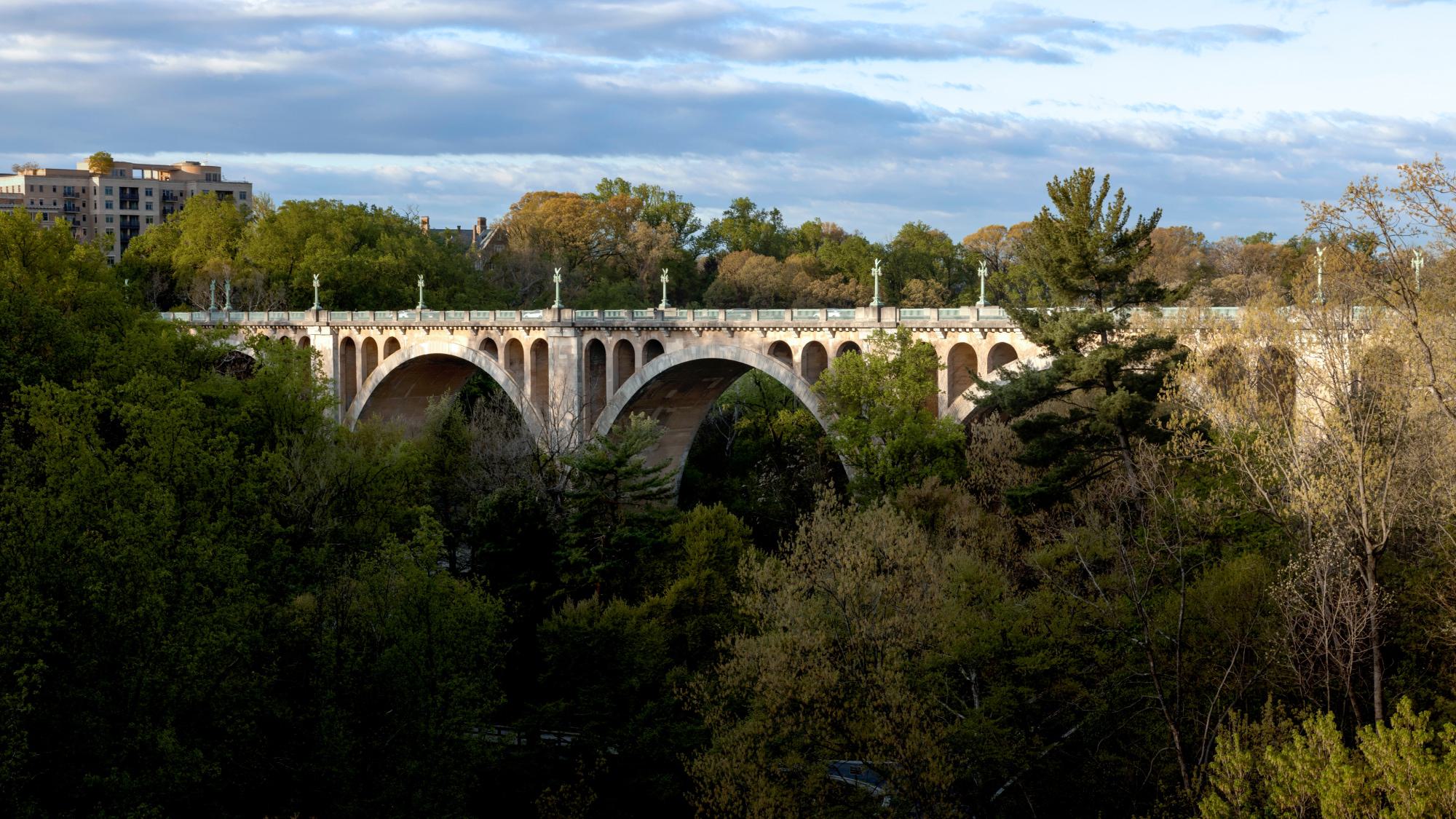 The historic Taft Bridge spans Rock Creek Park, its elegant arches rising above a dense canopy of spring foliage with apartment buildings visible in the distance under a partly cloudy sky.