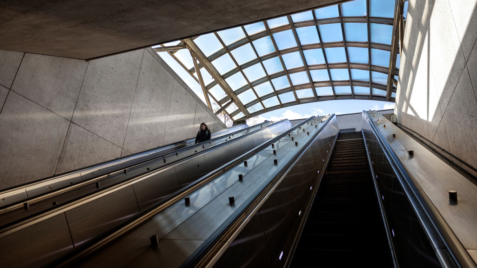 A person rides up the long escalators at the Woodley Park–Zoo/Adams Morgan Metro station, framed by the station’s curved glass canopy and angular concrete walls.