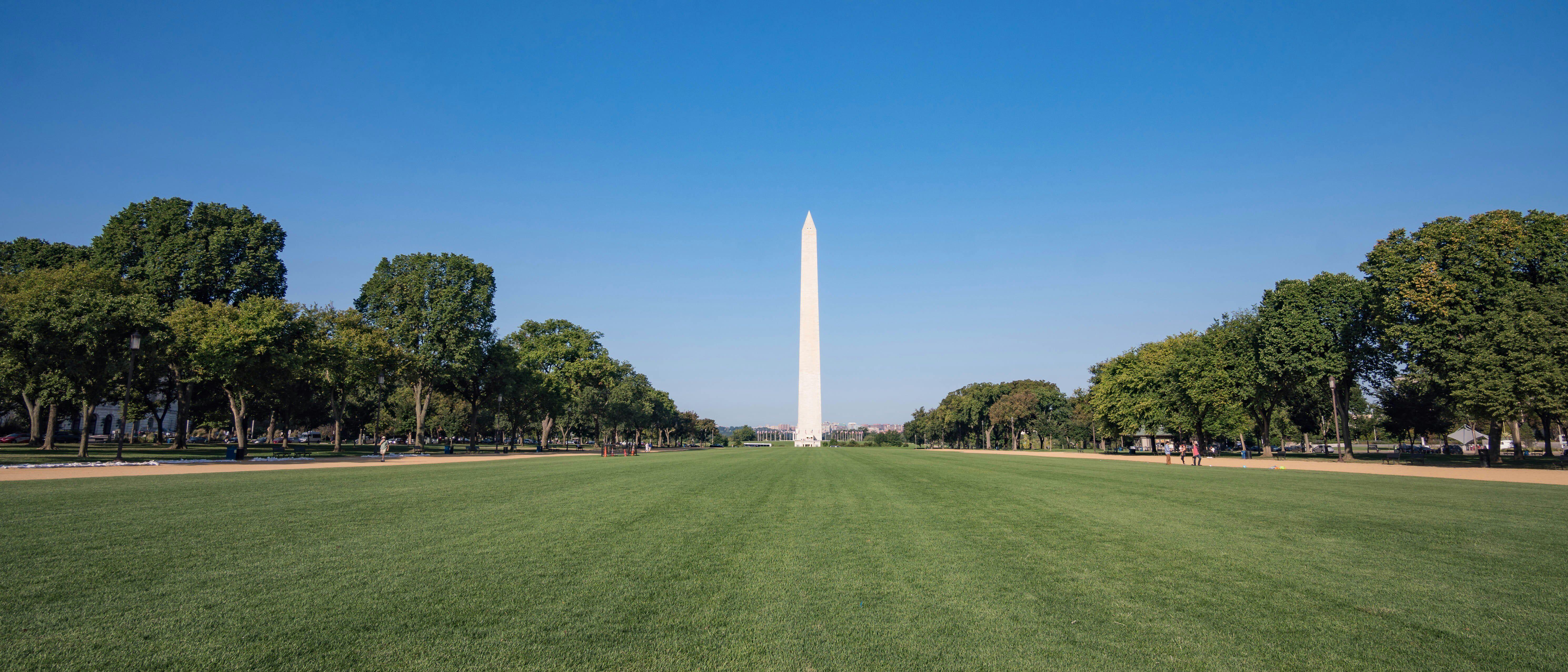 wide view of the National Mall in Washington, DC, with the Washington Monument centered in the distance, green lawns in the foreground, and trees lining both sides under a clear blue sky.