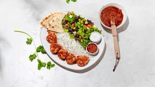 A plate of grilled chicken kabob pieces served over herbed basmati rice with a side of mixed green salad, warm pita wedges, and small bowls of red pepper sauce and tzatziki. Fresh cilantro stems and a bowl of extra red sauce with a brush sit beside the plate on a light textured surface.
