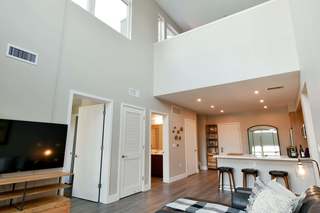 A modern loft-style apartment with double-height ceilings, tall upper-level windows, and a mezzanine overlooking the great room. The main level includes a kitchen with bar seating, light wood cabinetry, recessed lighting, and a living area with a sofa and TV. Doors to a bathroom and bedroom line the hallway beneath the loft.