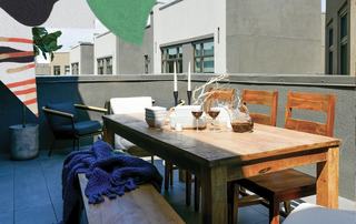 Outdoor balcony at Fourth Street East with a wooden dining table, bench seating, lounge chairs, and table settings, surrounded by modern building façades