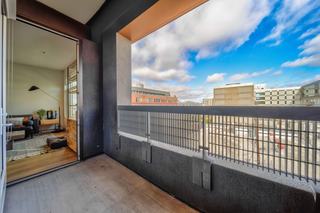 A spacious private balcony at Fourth Street East with dark gray walls and a metal railing, looking out over the urban cityscape of Jack London Square under a bright, cloudy sky. An open glass door reveals a glimpse of a modern, sunlit living room inside.