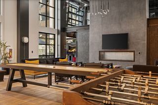 Design-forward clubhouse with game tables, tall black-framed windows, a charcoal tile fireplace wall, and lounge seating accented with yellow pillows.