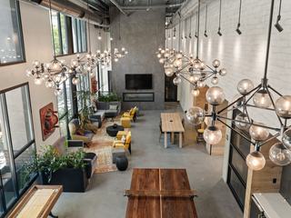 Coworking lounge with tall windows, sculptural chandeliers, lounge seating, a long communal table, shuffleboard and ping pong tables, greenery, and a modern fireplace wall.
