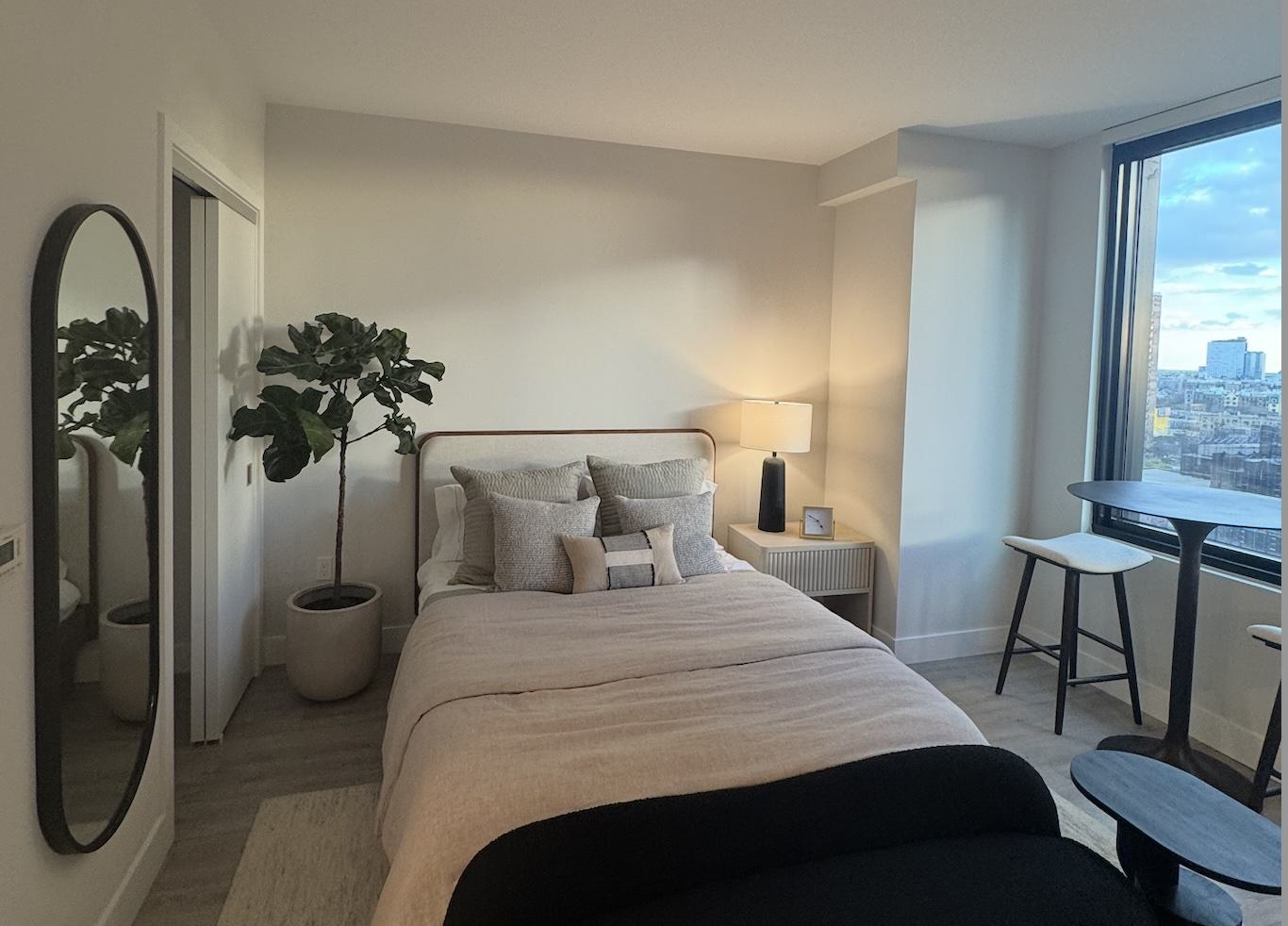 A bright, stylish bedroom featuring a neutral upholstered bed layered with pillows, a tall mirror, bedside table with lamp, large window with city views, and warm natural light.