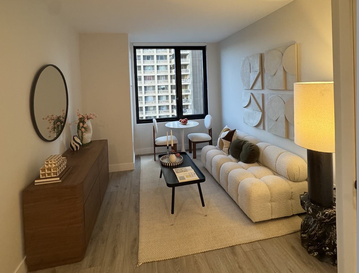 A modern furnished living room with a sculptural cream sofa, textured wall art, round dining set by a large window, wood sideboard with décor, and soft natural light.