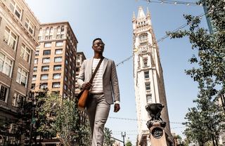 A man in a light gray suit walks confidently through a sunny downtown Oakland street, passing historic high-rise buildings and a sculptural pedestal, with string lights overhead and trees lining the urban walkway.