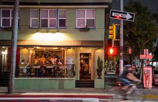 A lively Hillcrest street scene at night with diners gathered on a cozy restaurant patio, warm string lights glowing, and a cyclist passing through the crosswalk under a one-way sign.
