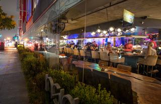 Nighttime view into a stylish Hillcrest restaurant and bar with glowing modern light fixtures, guests seated along a lively bar, and reflections of the busy street outside in the floor-to-ceiling windows.