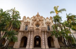 The ornate Spanish Colonial Revival façade of Casa del Prado in Balboa Park, featuring sculpted columns, intricate carvings, arched entryways, and tall palm trees framing the historic building.