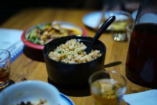 Black bowl of fried rice with green onions on a wooden table, surrounded by shared dishes, glasses, and a carafe, suggesting a casual group meal.