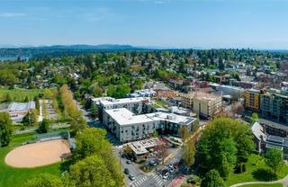 Aerial view of Columbia City in Seattle, featuring mid-rise apartments near the light rail station, surrounding tree-lined residential streets, neighborhood parks, and a dense green canopy extending across the Rainier Valley.