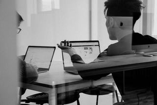 Two people working together at a table with laptops in a modern office setting, seen through glass.