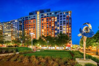 Evening view of modern mid-rise buildings with lit windows overlooking a landscaped park and abstract metal sculpture, capturing the urban streetscape and public art of Reston Town Center at dusk.