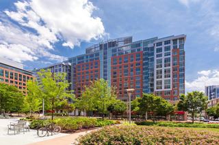 Daytime view of Reston Town Center featuring modern mid-rise buildings framed by trees, landscaped park space with seating and bike racks, and a bright, open plaza under a blue sky.