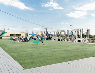 Rooftop lawn at Union Market District in Washington, DC, with people relaxing on artificial turf, picnic tables, string lights, and a food-and-drink container bar under a bright sky.
