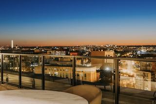 Rooftop balcony view over Washington, DC at dusk, with glass railing, city lights below, and the Washington Monument visible on the horizon against a fading sunset sky.