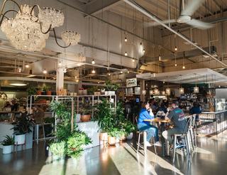 Interior of Union Market in Washington, DC, with hanging lights, lush plant displays, food stalls, and people dining at bar-height tables in a bright, open market hall.