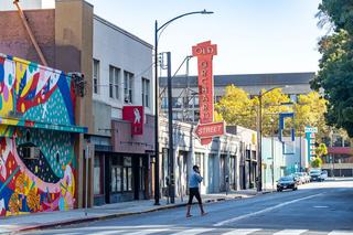 Colorful street scene on Old Orchard Street in downtown San Jose, featuring a vibrant mural on low-rise buildings, the vintage Old Orchard Street sign, and a pedestrian crossing a quiet urban roadway.