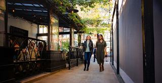 Two women walk through a cozy, plant-lined Ballard passageway beside an outdoor café, with string lights overhead and neighboring diners creating a lively yet relaxed neighborhood atmosphere.