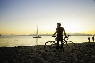 A cyclist stands on the sand at Golden Gardens Park at sunset, looking out over Puget Sound as sailboats pass and the Olympic Mountains line the horizon.