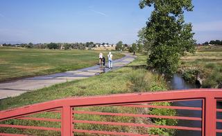 Paved walking path through a grassy open space, with people strolling alongside a narrow creek and a red pedestrian bridge in the foreground under a clear sky.