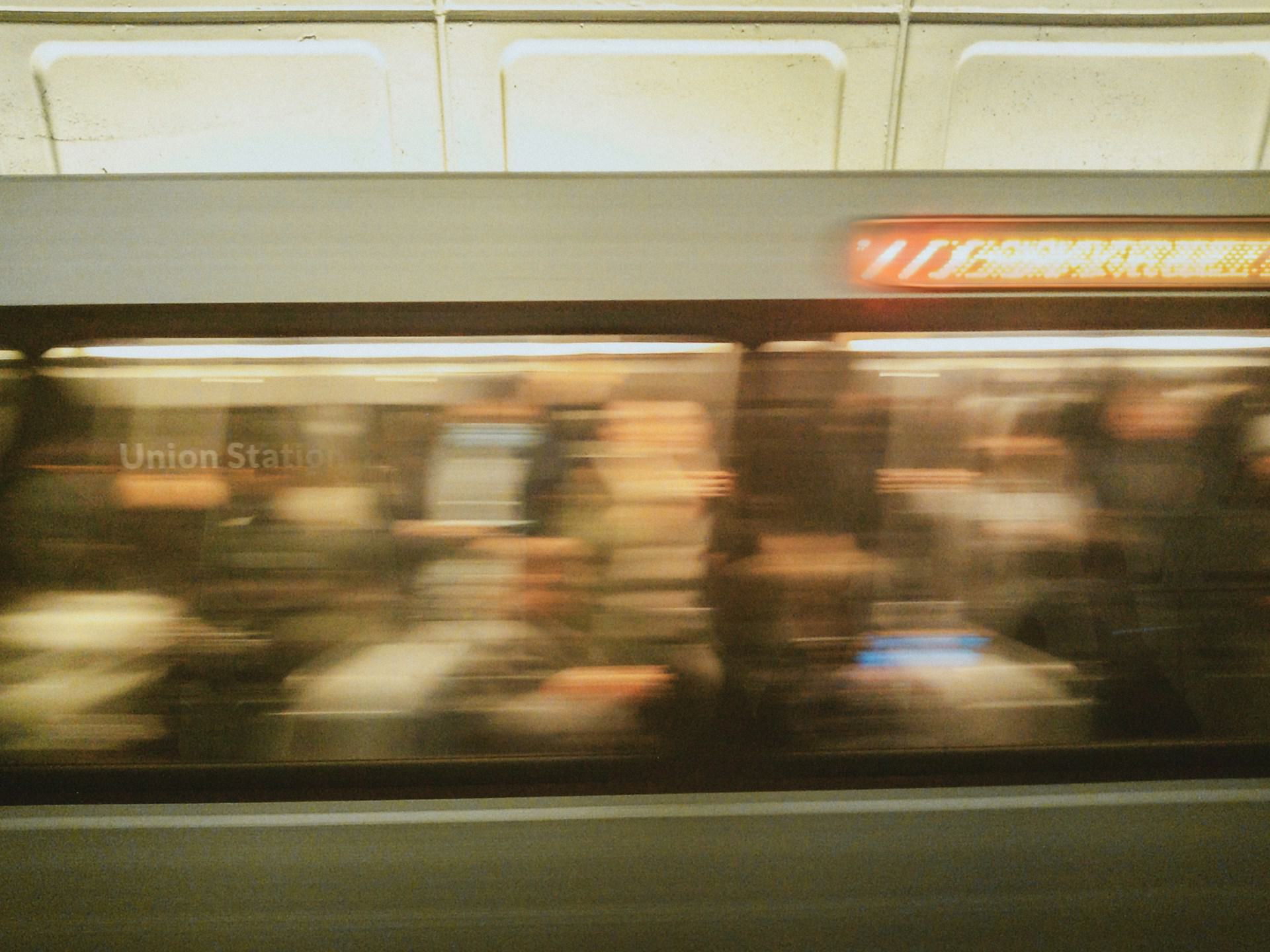 Blurred motion photo of a Washington Metro train arriving at a station, with passengers visible through the windows and an illuminated destination sign overhead.