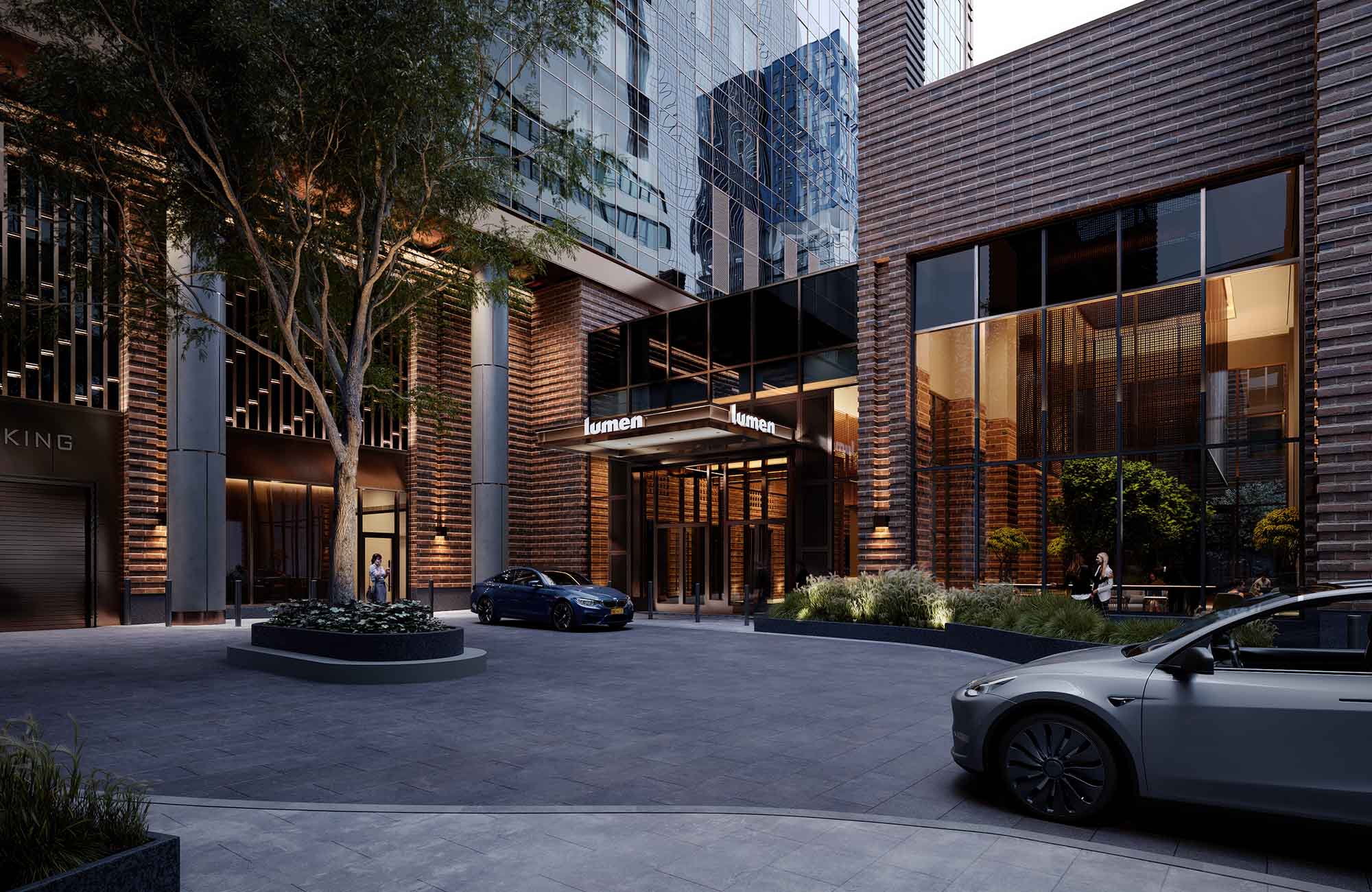 Street-level view of Lumen LIC apartments showing a modern brick-and-glass entrance with a covered porte cochère, landscaped planters, and cars pulling up at the lobby drop-off.