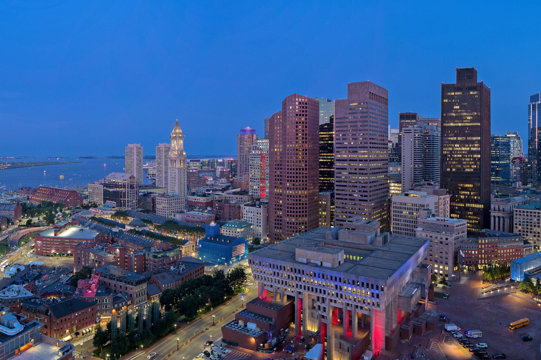 Downtown Boston skyline at dusk, with Financial District office towers rising behind Boston City Hall and Government Center Plaza, harbor visible in the distance and city streets lit below.