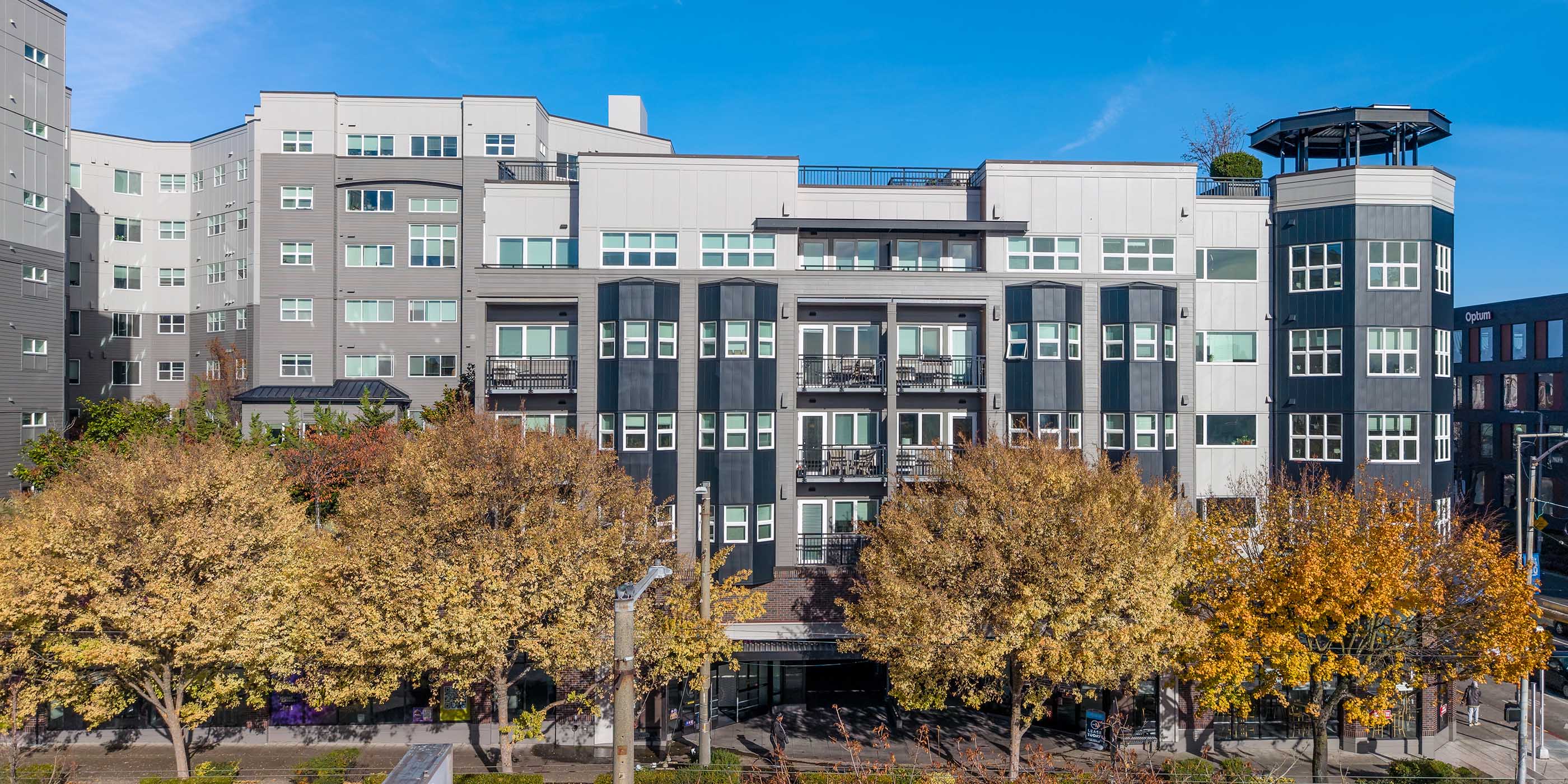 Modern apartment building in Seattle with gray and white façades, large windows, balconies, and street-level trees showing fall foliage.
