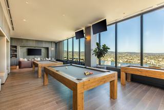 Games room with two full-size pool tables and a shuffleboard table set on wood floors, framed by floor-to-ceiling windows overlooking the Oakland hills and waterfront, with lounge seating and wall-mounted TVs in the background.