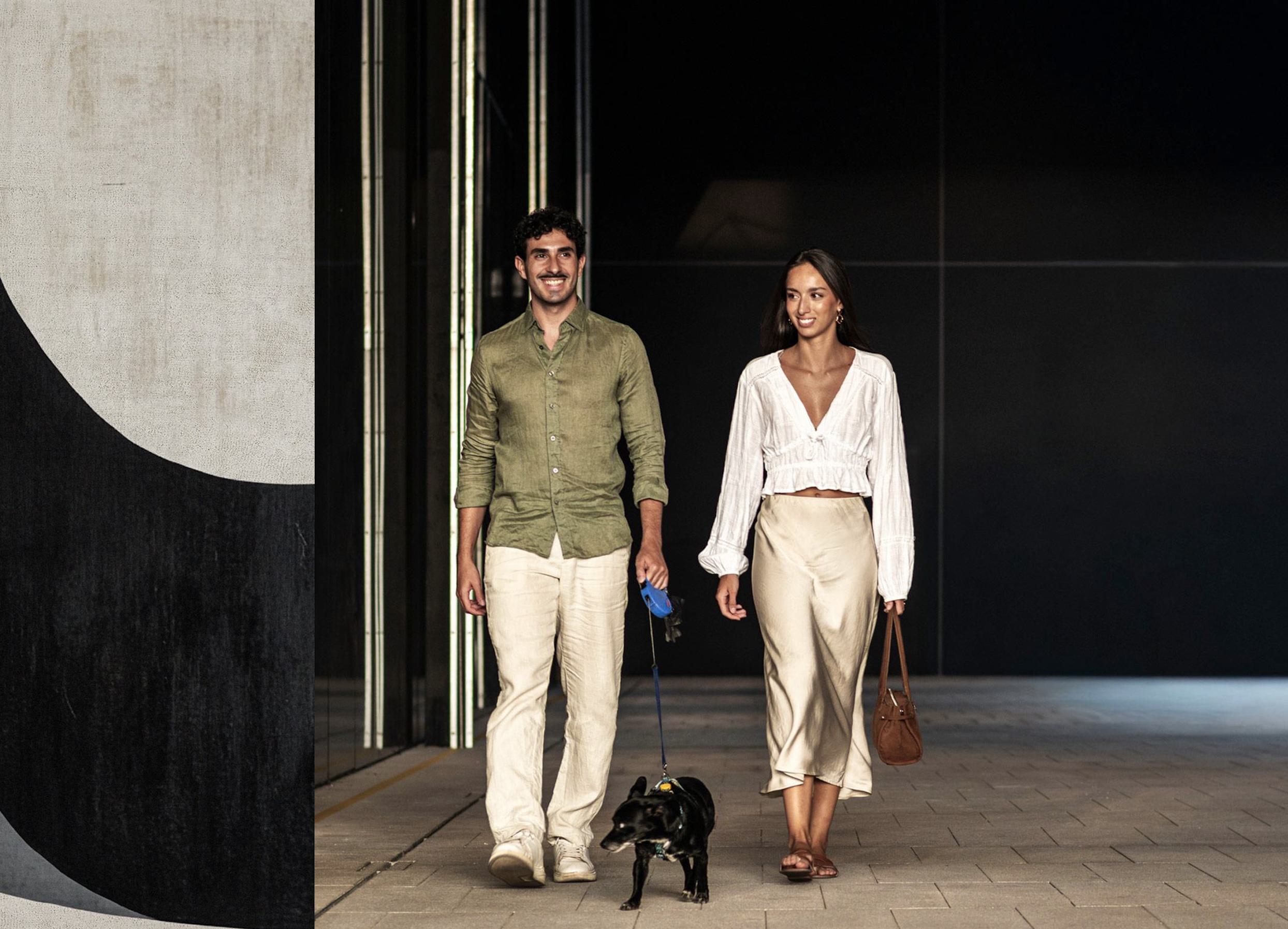Couple walking a small black dog on leash outside a modern high-rise building at dusk, contemporary façade and city setting in Tysons.