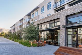 Modern apartment building at The Asher in Fremont with stone-and-stucco facade, large glass windows, balconies, landscaped walkway, outdoor seating, and views toward the rolling East Bay foothills.