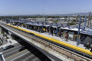 Elevated view of the Expo/Sepulveda Metro E Line station in Los Angeles, showing the platform’s wave-like canopy, arriving trains, and passengers boarding, with Sepulveda Boulevard below and the Century City skyline visible in the distance.