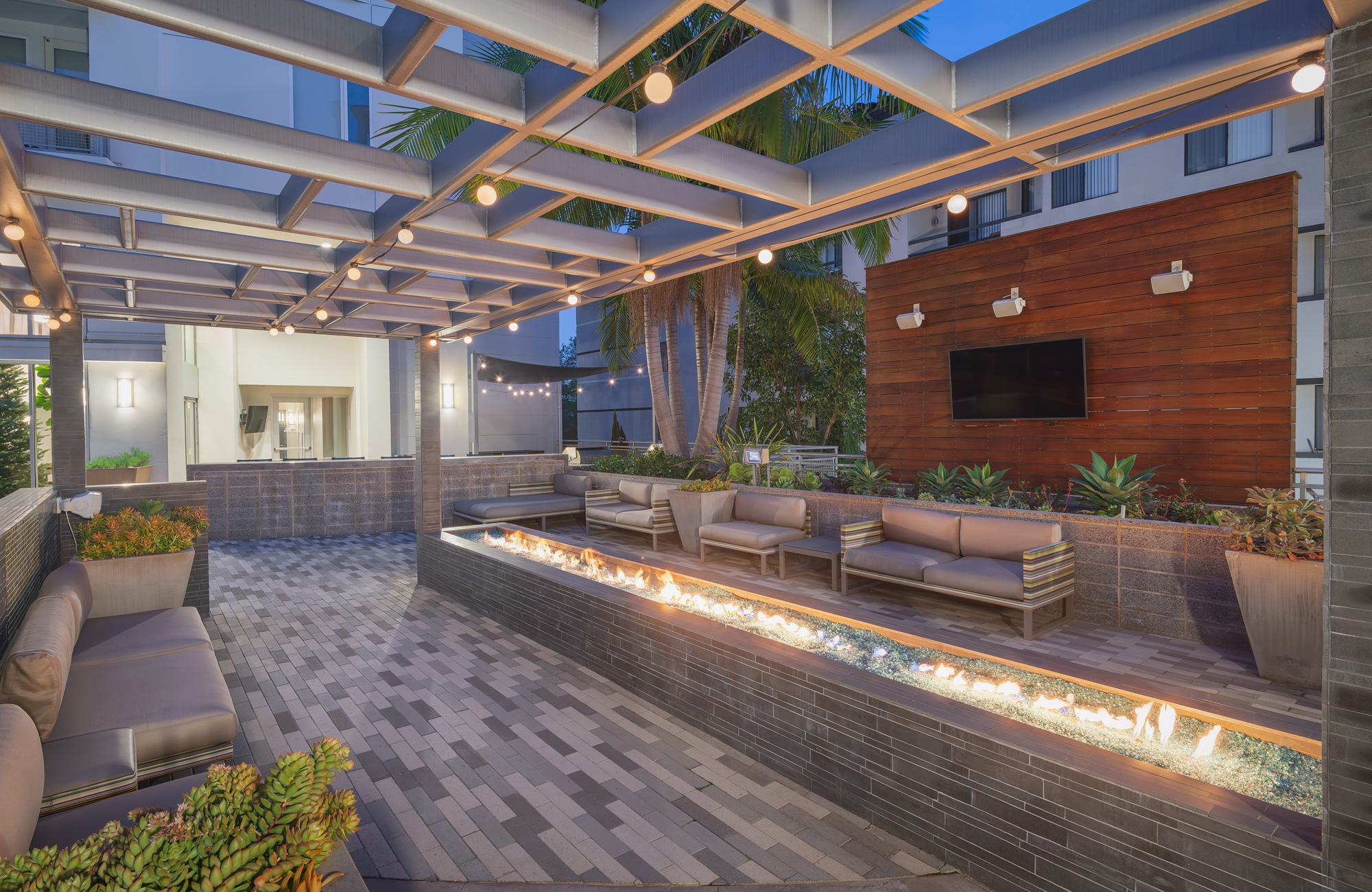 Covered outdoor lounge with a long linear fire feature, cushioned seating, pergola lighting, and a mounted TV in Desmond’s courtyard.