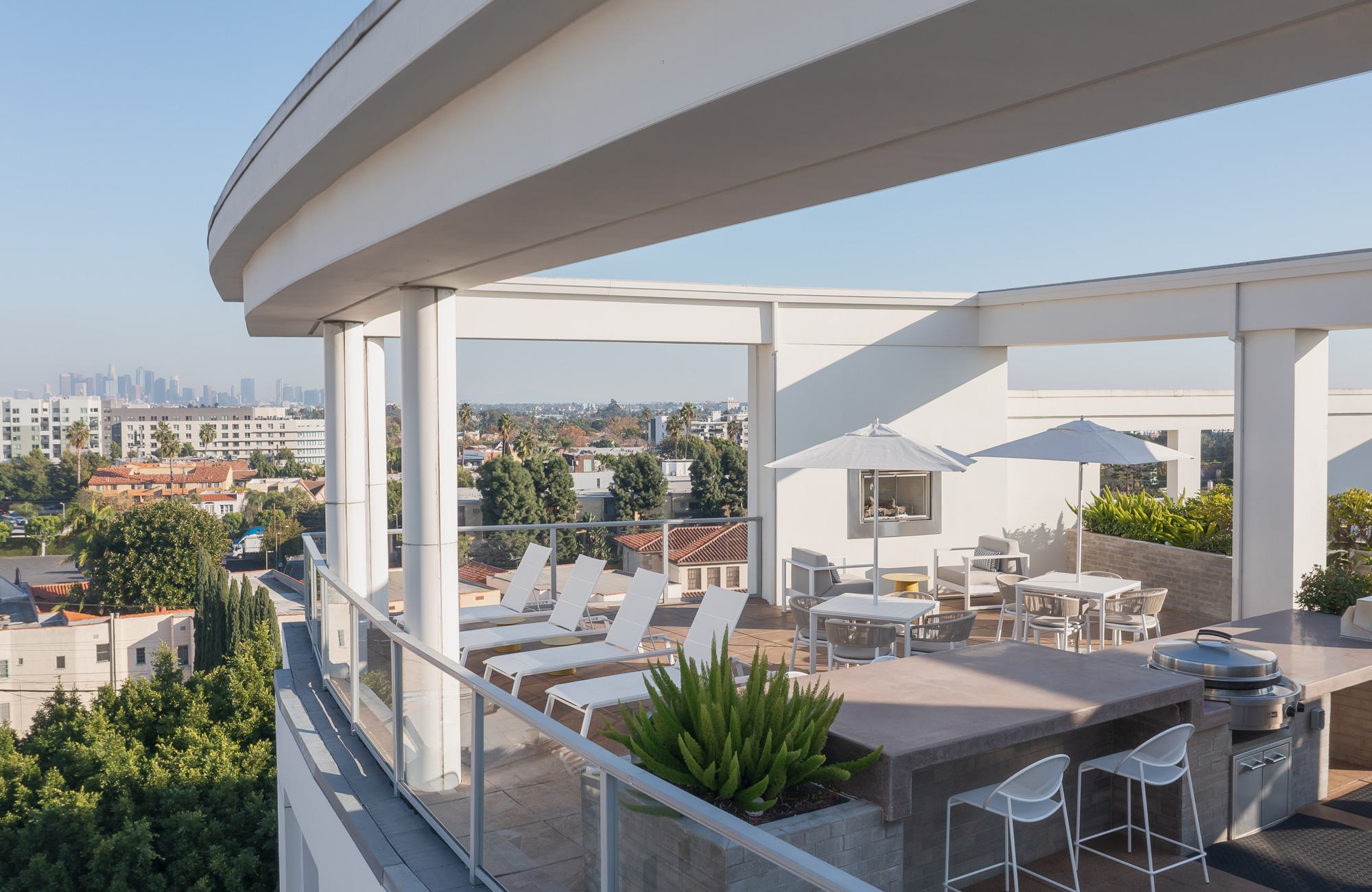 Rooftop lounge with white columns, built-in grills, dining tables, and chaise loungers overlooking the Miracle Mile neighborhood and the Downtown Los Angeles skyline.