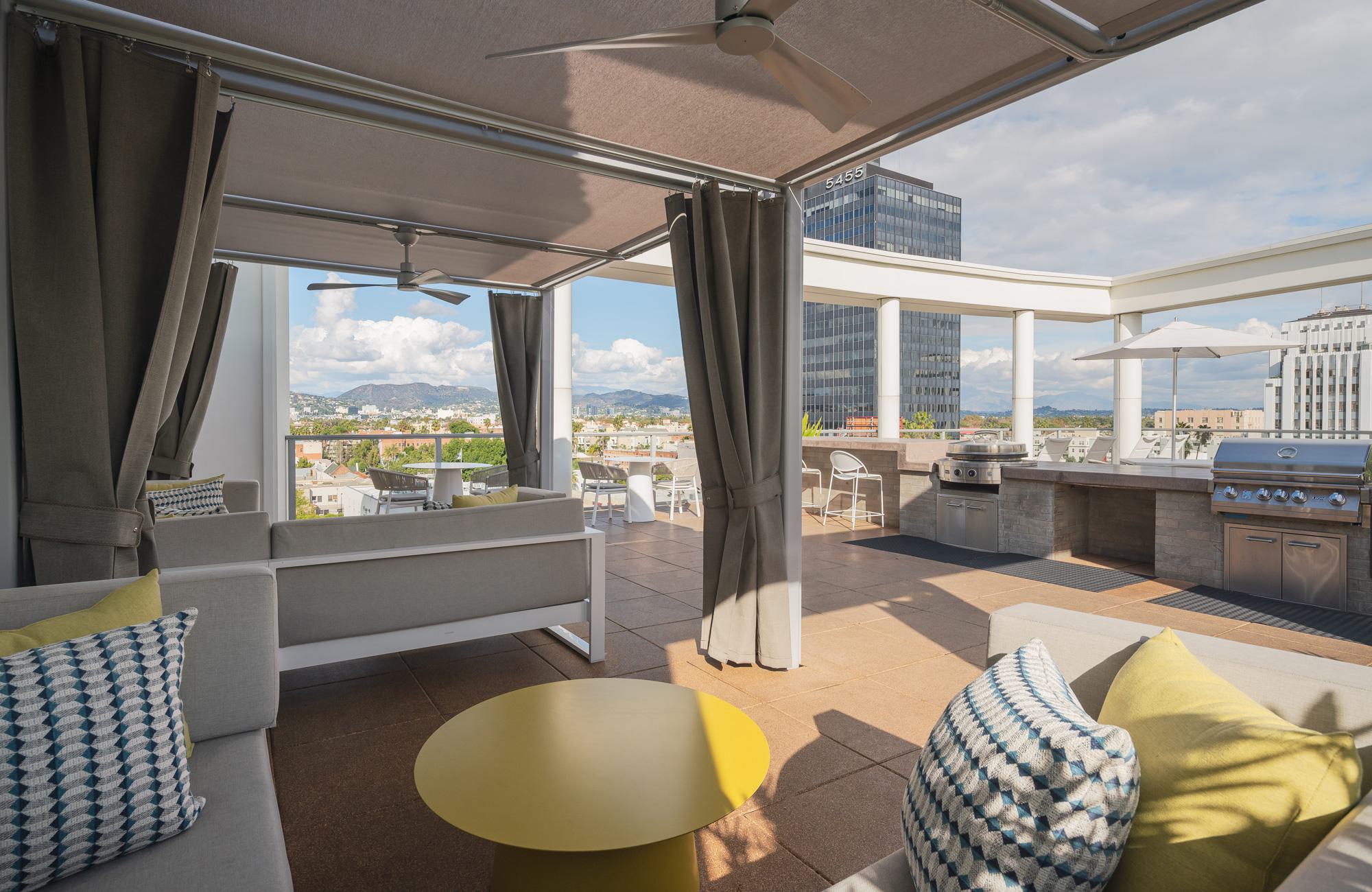 Covered rooftop cabanas with sofa seating, ceiling fans, and curtains, opening onto the Desmond rooftop with city and hill views in the distance.