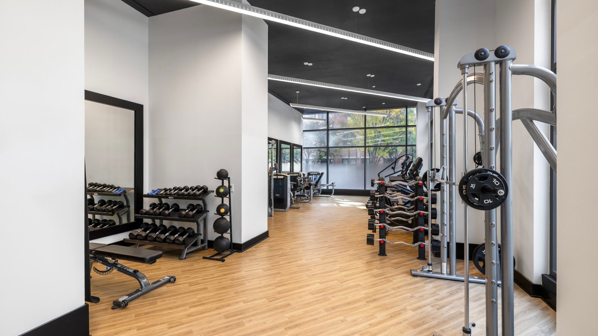 A bright, modern fitness center at Latitude featuring high ceilings, wood-style flooring, a dual-adjustable cable machine, and a weight area with dumbbells and medicine balls near large windows with privacy film.
