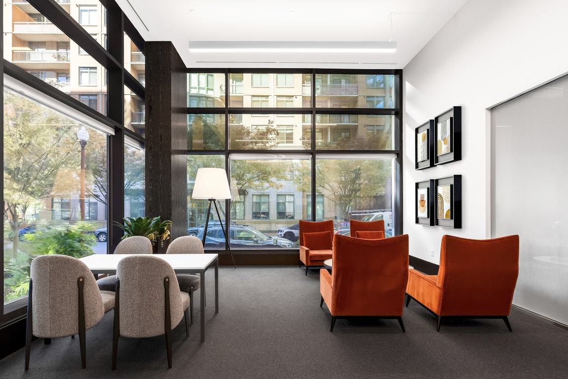 A bright corner lounge area featuring vibrant orange armchairs, a small meeting table with grey chairs, framed modern art, and wrap-around floor-to-ceiling windows.