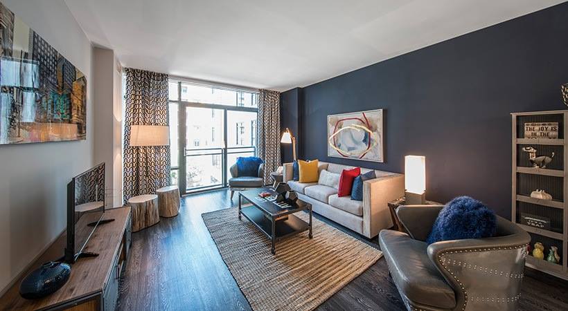 Sunlit living room with floor-to-ceiling glass doors opening to a private balcony, featuring a modern sofa, accent chairs, and city views beyond.