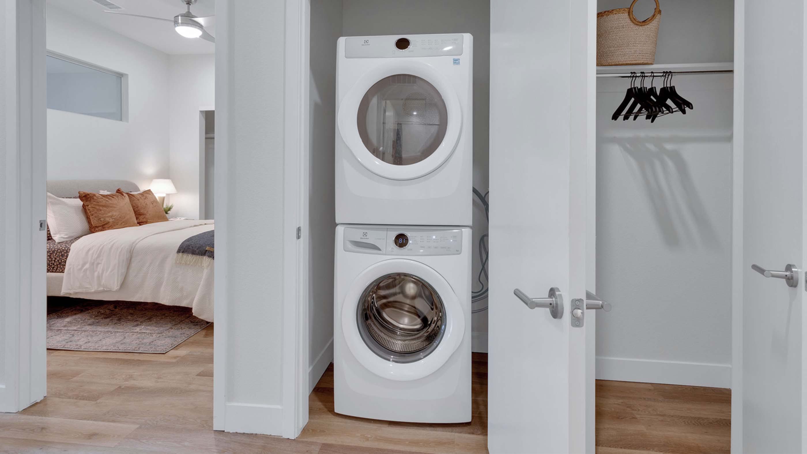 Stacked in-home washer and dryer tucked into a hallway closet at Latitude, with a bedroom visible beyond and built-in storage to the side.