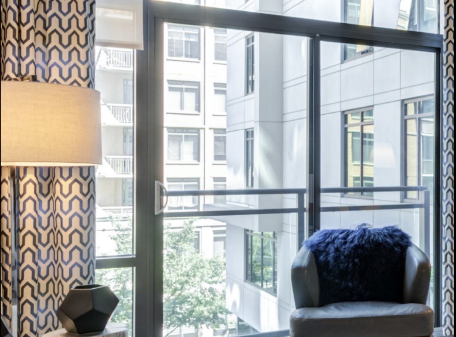 Apartment living room with floor-to-ceiling windows and a glass door to the balcony, filling the space with sunlight and framing views of the surrounding buildings.