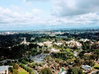 Aerial view of Balboa Park in San Diego, showing the museum complex, California Tower, tree-filled canyons, and surrounding neighborhoods near Hillcrest under a cloudy sky.
