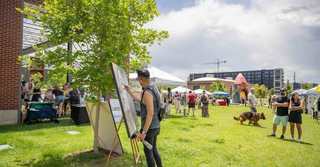 Artist painting at an outdoor art market on the lawn at RiNo ArtPark in Denver, with visitors, tents, public art sculptures, and modern buildings in the background.
