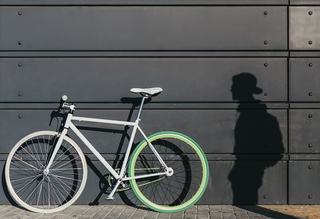 White minimalist bike with a bright green rear rim leaning against a dark metal wall, with a person’s shadow cast beside it on the sidewalk.