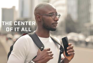 A man wearing glasses and a white t-shirt stands outdoors in a city setting, holding a smartphone and adjusting his backpack straps. Skyscrapers blur in the background, and bold text on the image reads, “THE CENTER OF IT ALL.”