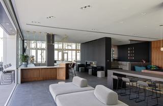 Spacious resident clubhouse with floor-to-ceiling sliding glass doors, dark tile flooring, wood-paneled bar, brass pendant lighting, built-in fireplace, and modern lounge seating areas.